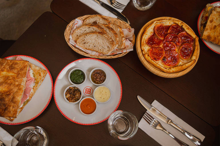A foto mostra uma mesa com diversos pratos brancos com bordas vermelhas que acomodam um sanduíche, cinco potinhos com antepastos coloridos, uma torta de frutas e uma cesta com fatias de pão