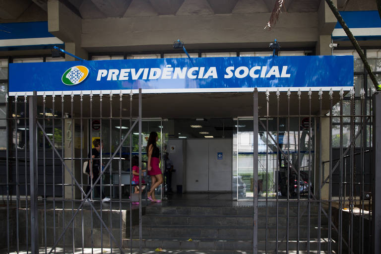 A imagem mostra a entrada de uma agência da Previdência Social no Brasil, com um portão de grade fechado e uma placa azul com o logotipo e o nome "Previdência Social" em destaque. Pessoas podem ser vistas ao fundo, dentro da estrutura, sugerindo atividade no local.
