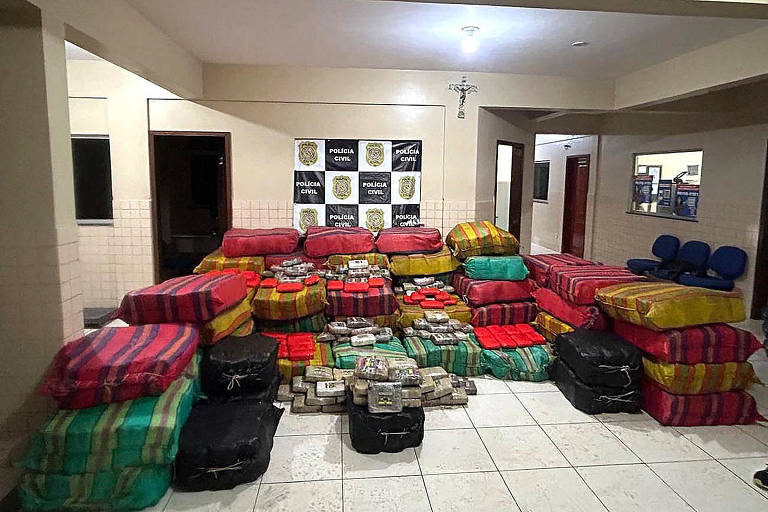 Uma sala com paredes claras e uma placa da Polícia Civil ao fundo, com cargas de drogas dentro de pacotes e bolsas coloridos empilhados no chão 