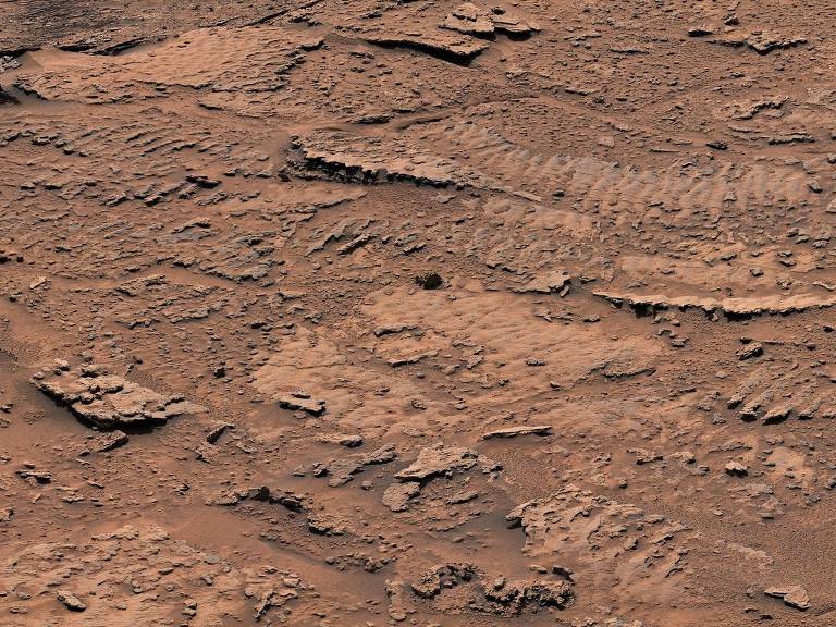 A imagem mostra uma superfície rochosa com várias formações e texturas. O terreno é predominantemente marrom-avermelhado, típico da superfície de Marte. Há várias rochas de diferentes tamanhos e formas espalhadas pelo solo, com algumas áreas apresentando padrões de erosão e fissuras.
