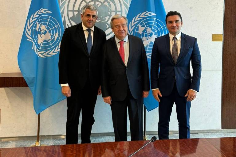 Ministro Celso Sabino, secretário-geral da ONU e  secretário-geral da ONU Turismo sorriem em frente à bandeira da ONU
