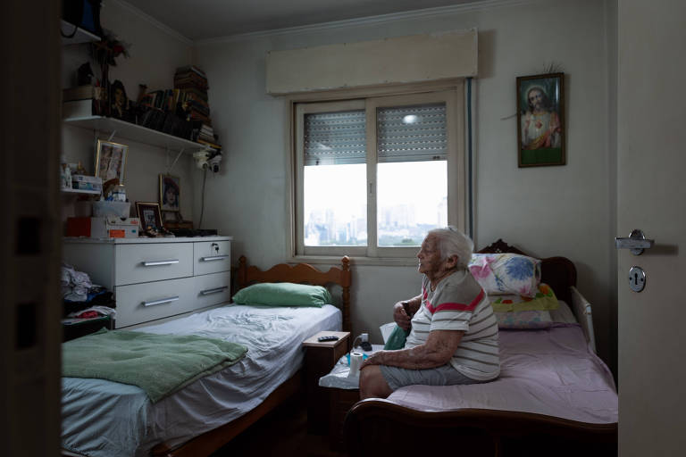 Na imagem é possível observar Martha sentada numa cama com lençol cor-de-rosa. Ao fundo, tem uma janela com vista para São Paulo. No mesmo ambiente outros móveis como uma cômoda e uma outra cama estão alocados.