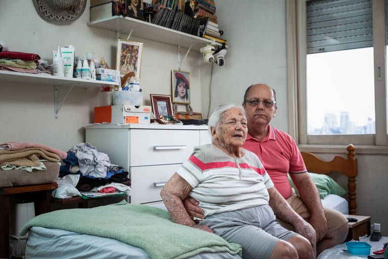 Idosa de 102 anos sentada na cama ao lado do filho