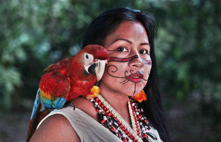 Em foto colorida, a cantora indígena Denizia Araújo Peres posa para a câmera com uma arara pousada em seu ombro