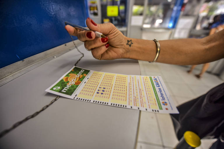 Imagem fechada mostra uma mão segurando uma caneta sobre um volante da Mega-Sena com números marcados