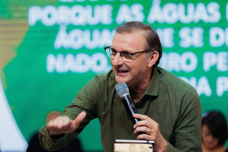 Jucélio de Souza, um homem branco com cabelo liso, curto e castanho claro usa óculos e fala ao microfone