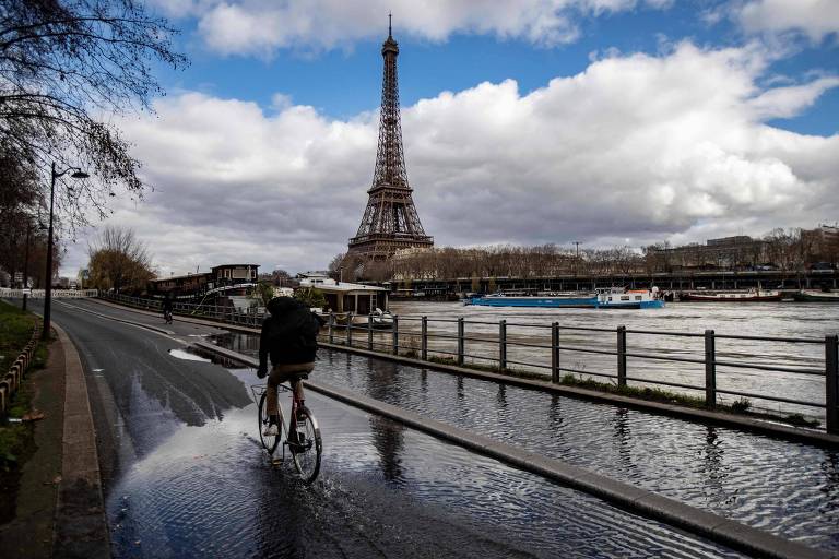 Ciclista em margem do rio Sena, em Paris; há uma poça de água refletindo o céu; ao fundo está a torre eiffel