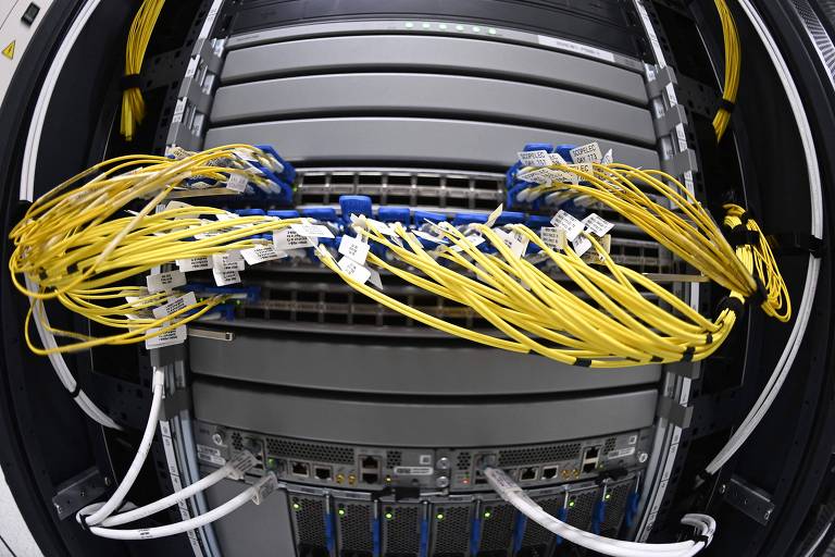 A imagem mostra um data center, com um servidor de rede com vários cabos conectados. Os cabos são predominantemente amarelos e estão organizados em fileiras, conectados a portas de rede no servidor. A estrutura do servidor é metálica e possui várias camadas de conexões.