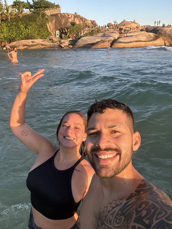 selfie de casal em praia, com mulher fazendo V com os dedos