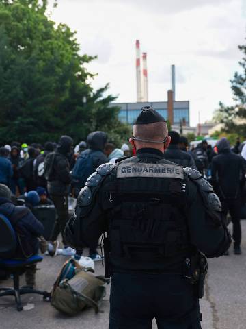A French gendarme stands guard next to migrants waiting with their belongings to be registered by associations members to get to another location, during the evacuation of France's biggest squat, which has housed up to 450 migrants, most of them legal migrants according to associations - in the southern suburbs of Paris in Vitry-sur-Seine on April 17, 2024, 100 days before the Olympic Games. The operation, which had been expected for several days, prompted many of the homeless who had taken refuge in this disused factory in Vitry-sur-Seine (Val-de-Marne) to leave before the arrival of the large numbers of police deployed to dislodge them. (Photo by Emmanuel Dunand / AFP)