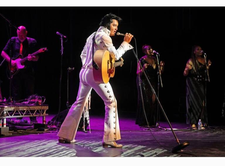 Em foto colorida, o performer britânico Ben Portsmouth aparece vestido de Elvis Presley cantando em um palco