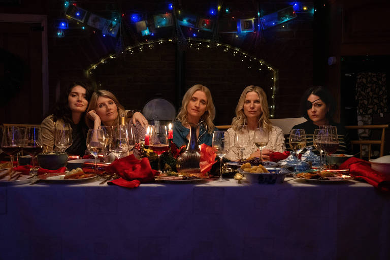 Cinco mulheres estão sentadas lado a lado a uma mesa de jantar, cheia de copos, pratos com comida e velas. Todas elas olham para a câmera de um jeito misterioso