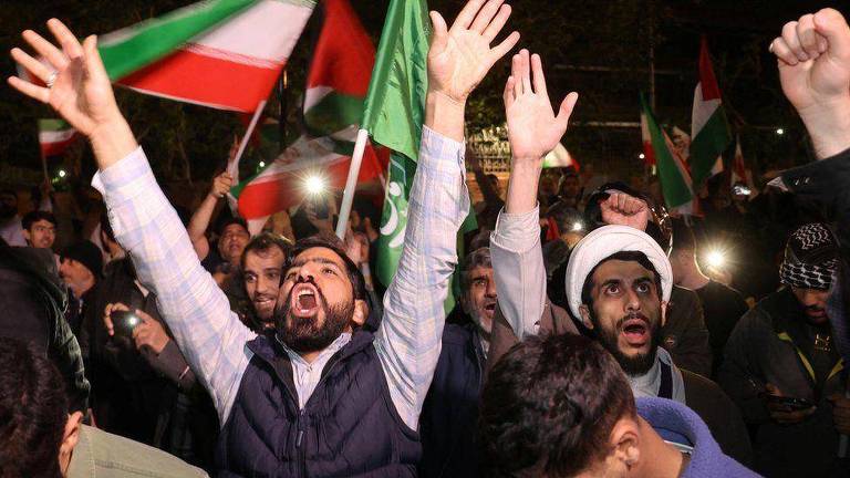 Homens com braços estendidos em meio à multidão e bandeiras do Irã ao fundo
