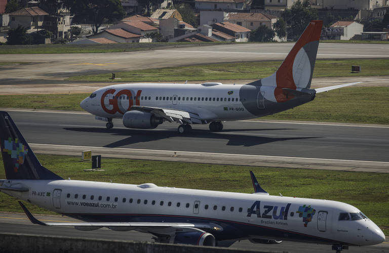 Dois aviões posicionados em uma pista de aeroporto. Um avião tem detalhes em azul e tem o logo da companhia aérea Azul e o outro tem detalhes em laranja e o símbolo da companhia aérea Gol.