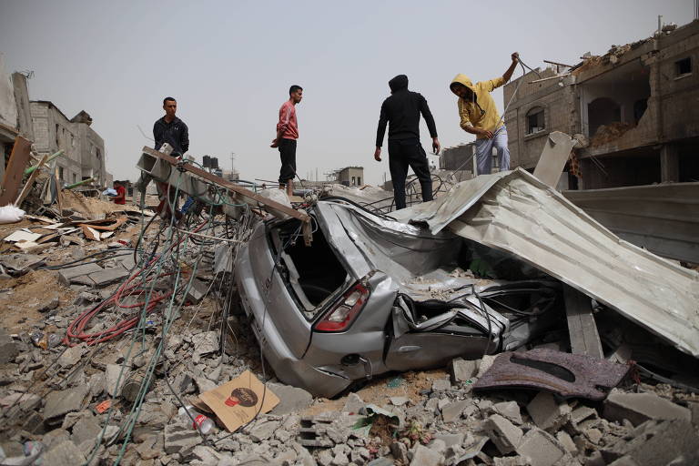 Pessoas de pé sobre destroços de concreto e metal, com carro destruído em primeiro plano 