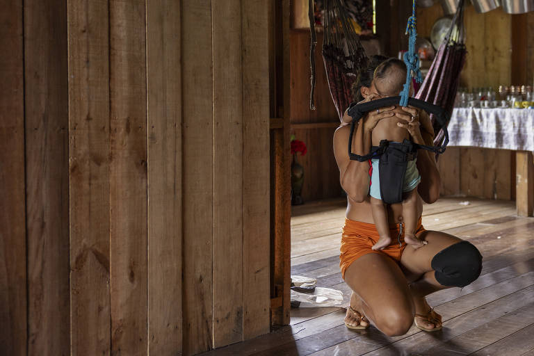 Foto de uma jovem agachada com um bebê do costas sobre suas pernas. A cabeça do bebê encobre o rosto da menina. As paredes e chão do local são feitos de tábua e o bebê está envltou em cordas, como uma rede suspensa.