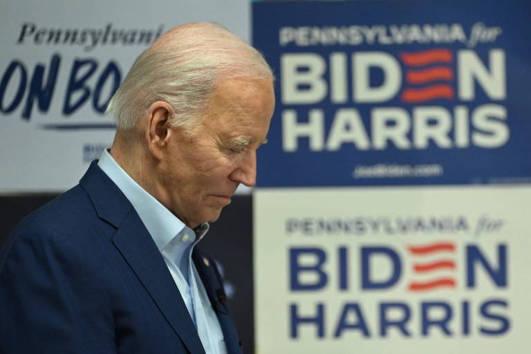 Joe Biden com a cabeça baixa e olhos fechados; ao fundo, emblemas da campanha democrata.