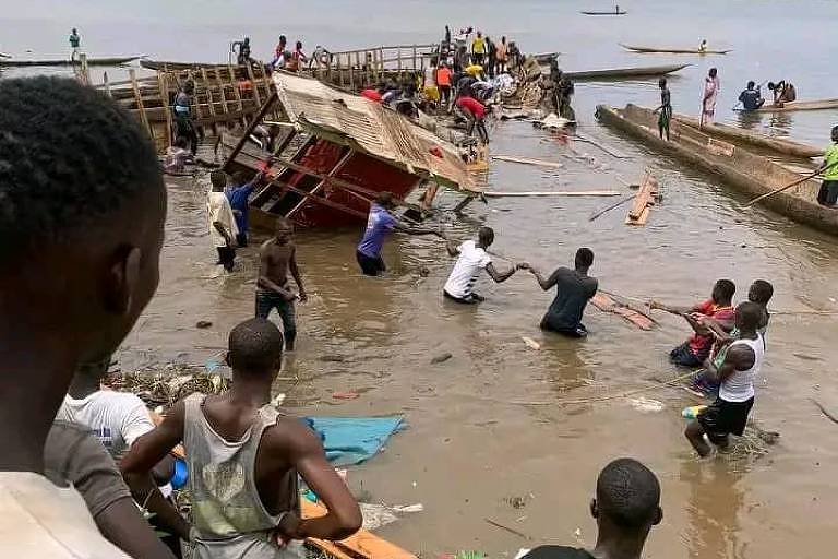 Foto mostra um rio, com destaque para um barco de madeira naufragado; as estruturas do barco estão quebradas e, sobre elas, pessoas se equilibram para resgatar vítimas do acidente