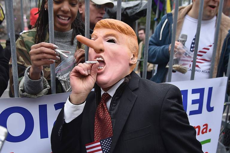 Homem vestido com terno e com uma máscara do rosto de Donald Trump (rosto branco, cabelo loiro e nariz grande) fuma um cigarro de maconha