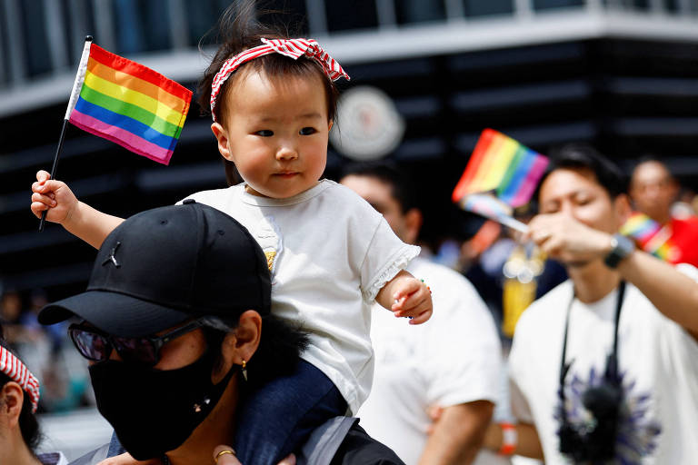 Criança sobre os ombros de adulto segura bandeira arco-íris