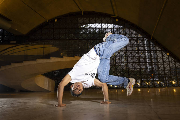 A imagem mostra um dançarino de breaking, realizando um movimento acrobático, apoiado com as mãos e com as pernas no ar. Ele está vestindo uma camiseta branca e calça jeans