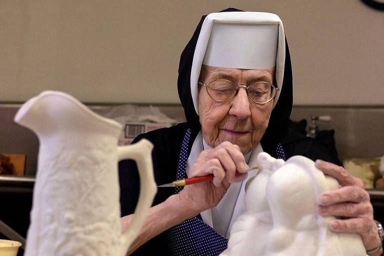 freira trajada com hábito faz escultura, há outra peça sobre a mesa, uma jarra 