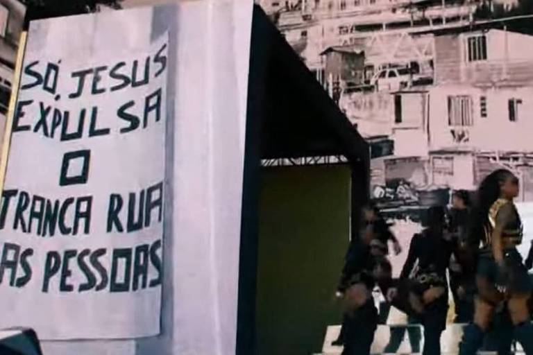 Projeção de vídeo exibe cartaz com mensagem "só jesus expulsa o tranca rua das pessoas". ela está do lado direito da imagem, no palco, junto com dançarinos