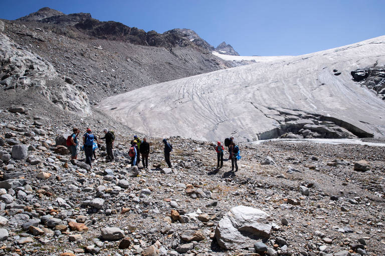 Pessoas em montanha com areia e pedras no chão; há gelo apenas no alto de uma montanha ao fundo