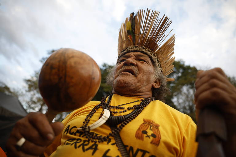 Indígena usa camisa amarela e colares com dentes de animais. Ele segura em uma das mãos um maracá. Na cabeça o ancião usa um cocar de palha. 