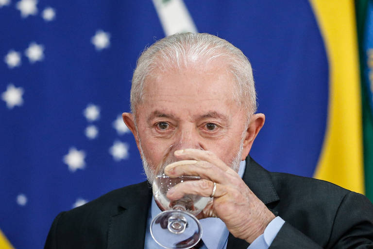 Lula bebendo água, de terno e gravata, em foto colorida. No fundo, a bandeira do Brasil