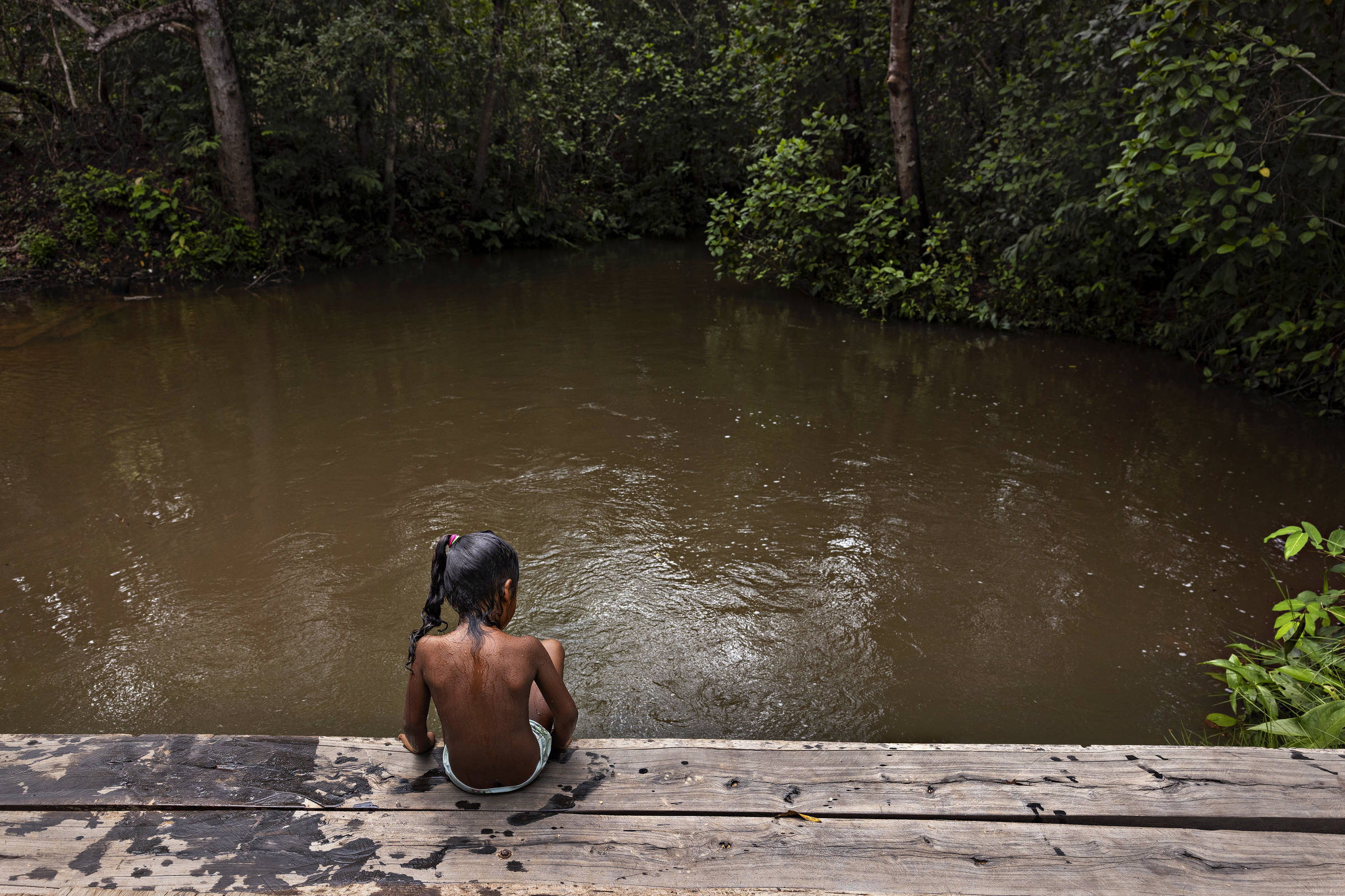 Menina negra, de costas, sentada em ponte de madeira sobre rio de água barrenta cerca por árvores 