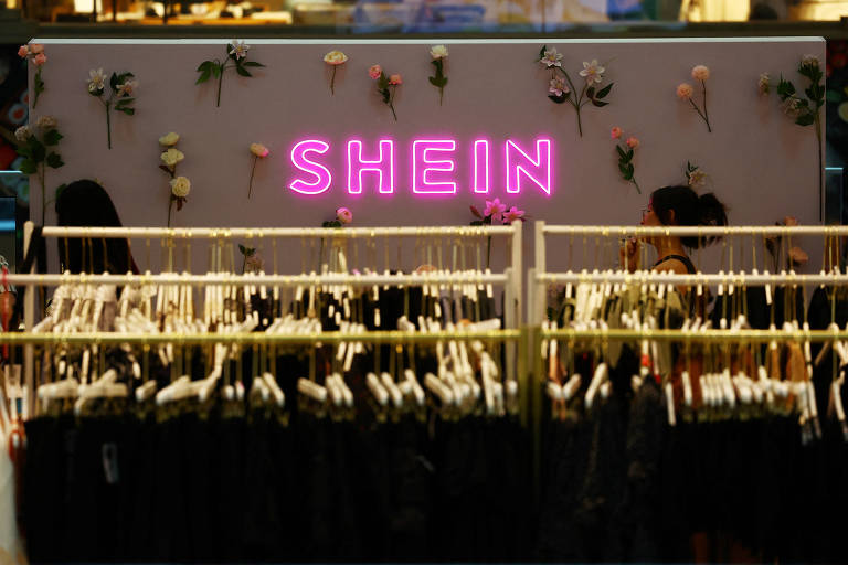 A imagem mostra o interior de uma loja da Shein com o logotipo da marca em neon rosa destacando-se no fundo decorado com plantas. Na frente, cabides alinhados exibem uma variedade de roupas, sugerindo um ambiente de compras moderno e acessível.