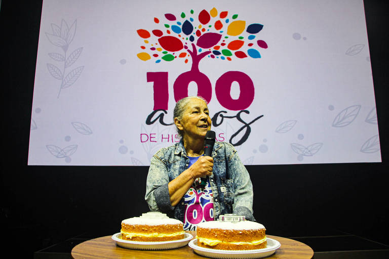 Mulher sentada, falando ao microfone. Na mesa à frente dela há dois bolos; ao fundo, uma faixa que diz "100 anos".