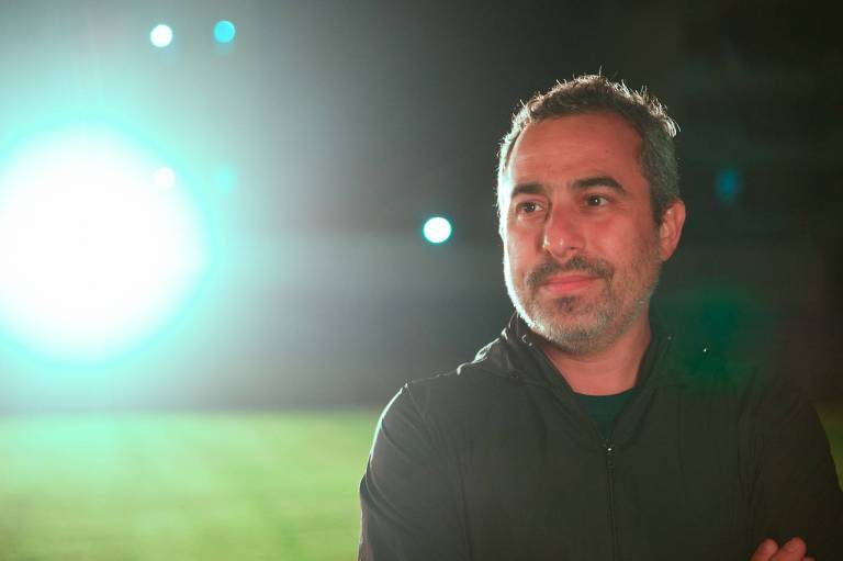 Foto do diretor Pedro Asbeg com roupa preta e um gramado de futebol ao fundo