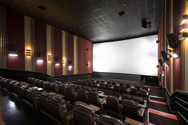 Sala de cinema vazia e com luzes acesas; telão está sem projeção ligada e paredes são revestidas nas cores vermelho escuro e bege