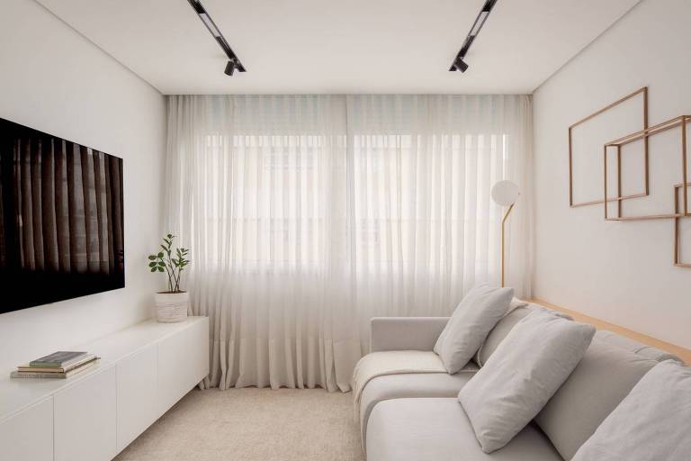 Sala em tons claros com sofá de um lado e tv no outro; e janela na lateral
