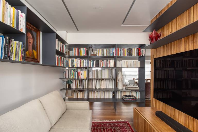 Sala com sofá claro escostado na parede e na frente dele uma TV em um painel de madeira; ao fundo, uma estante de livros