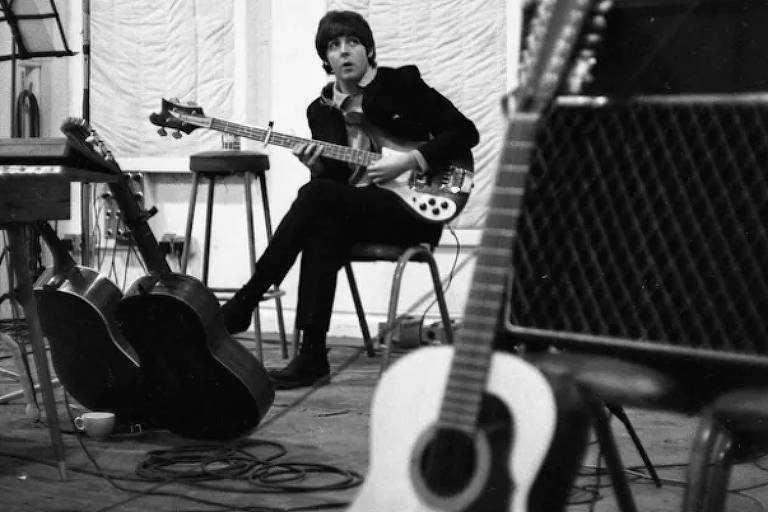 Foto em preto e branco de John Lenonn tocando guitarra. Um violão está à sua frente