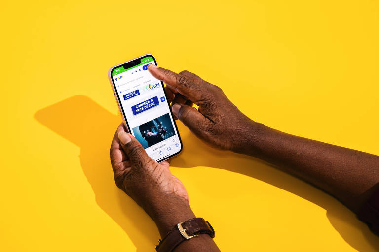 Sobre fundo amarelo, duas mãos seguram um celular. Na tela, podemos ver o App oficial do FGTS.
