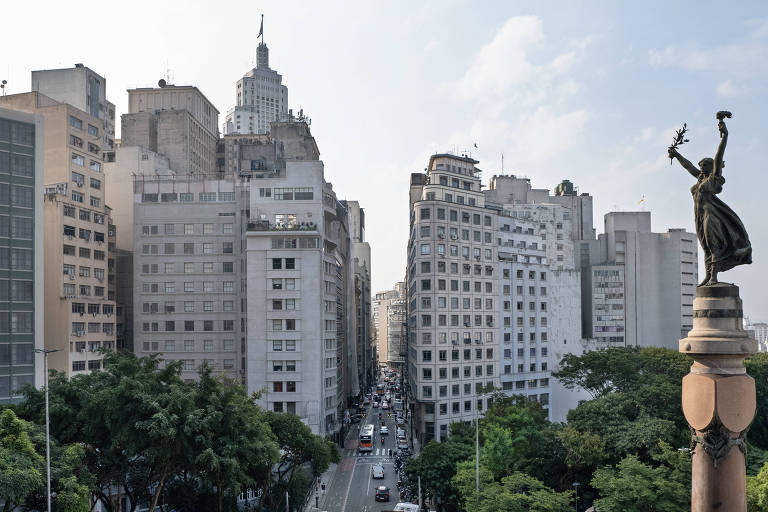 A estátua escura, de bronze, é uma figura feminina que usa vestido esvoaçante e ergue os dois braços, sendo possível ver que tem uma tocha em uma das mãos. Ela está sobre um alto pedestal e, ao fundo, é possível ver uma rua que está entre grandes prédios 