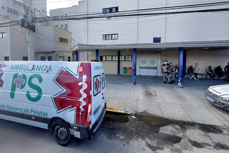 Uma ambulância branca está parada em frente a um prédio branco com vigas azuis. No veículo está escrito ambulância Pronto-Socorro de Pelotas.