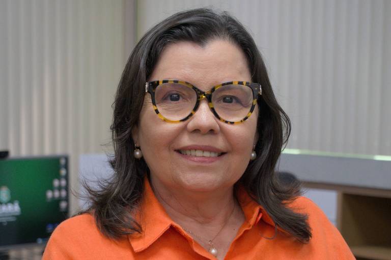 Sandra é uma mulher branca de cabelos escuros e curtos e olhos escuros. Na imagem, ela veste uma camisa de botão laranja e óculos de armação.