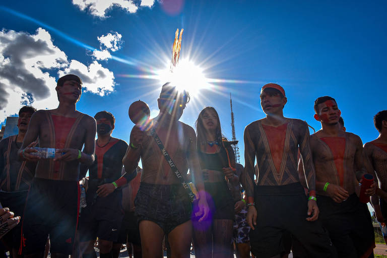 Indígenas com corpos pintados de preto e vermelho com cocares na cabeça, ao fundo uma forte luz do sol e um céu azul