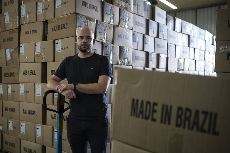 Foto mostra Israel em um estoque repleto de caixas com os dizeres "Made in Brazil"; Israel é um homem branco, careca e com barba castanha, e veste camiseta e calça pretas