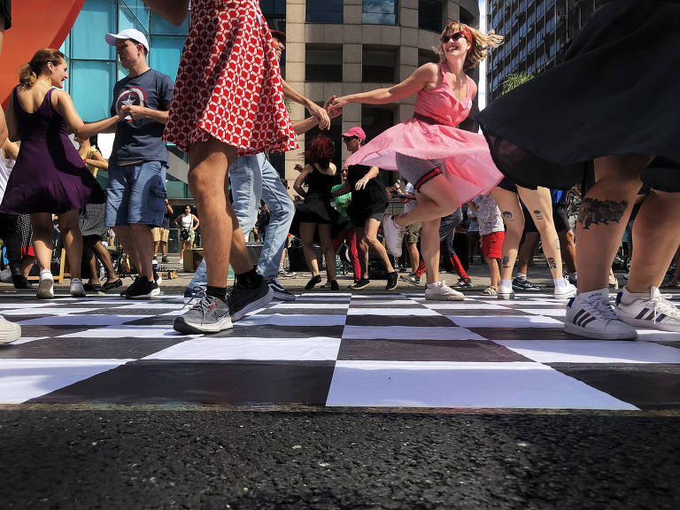 Imagem geral mostra pessoas dançando em pista com o chão quadriculado de preto e branco em ambiente externo.