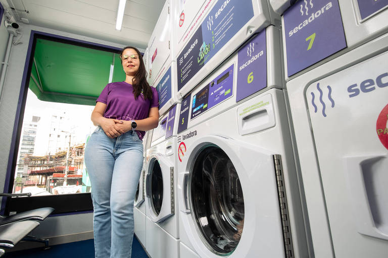 Foto mostra Heidi em pé， apoiada ao lado das máquinas secadoras de sua lavanderia; Heidi é uma mulher branca com cabelos longos e castanhos; ela veste camiseta roxa， calça jeans azul-clara e usa óculos de grau com armação marrom