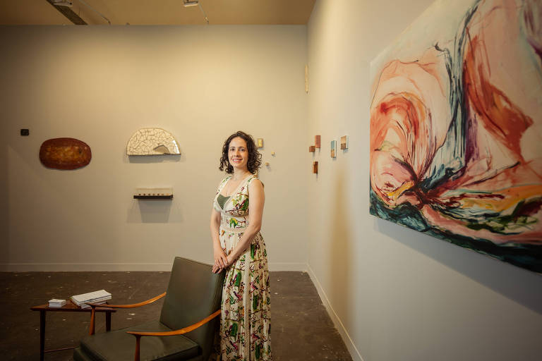 Retrato de Vanessa, uma mulher branca, com cabelos ondulados e castanhos na altura do ombro, em uma galeria de arte; Vanessa usa um vestido florido e está em pé, com as mãos apoiadas em uma cadeira