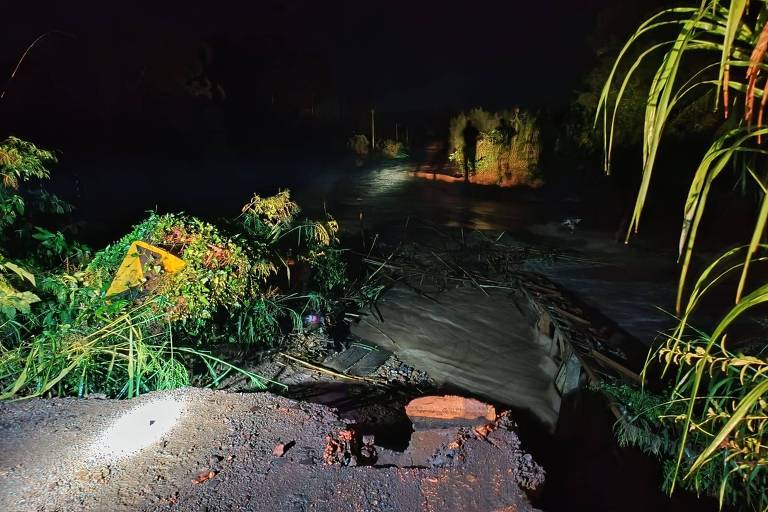 Na foto, tirada à noite, aparece uma estrada com um trecho desmoronado. Ao fundo há pessoas com capas de chuva, e vegetação nas laterais