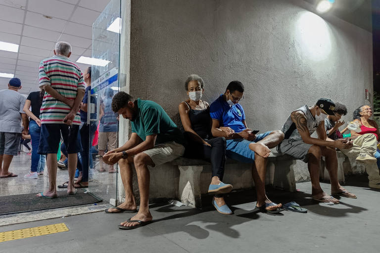 A imagem mostra várias pessoas sentadas em bancos de concreto ao lado de uma entrada iluminada, que parece ser de um hospital ou unidade de saúde. Algumas pessoas estão com expressão de cansaço ou desconforto, e uma delas usa máscara facial. Outras estão em pé, formando uma fila, sugerindo espera para atendimento. O ambiente é simples, com iluminação artificial, e o local parece movimentado, típico de um pronto-socorro ou unidade médica pública.






