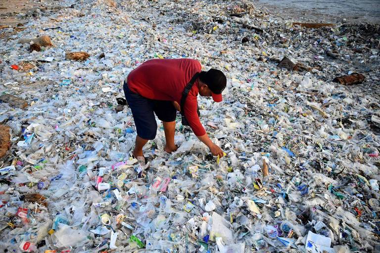 Inclinado, homem junta pedaços de plástico imerso em uma poluição que vai até os seus joelhos